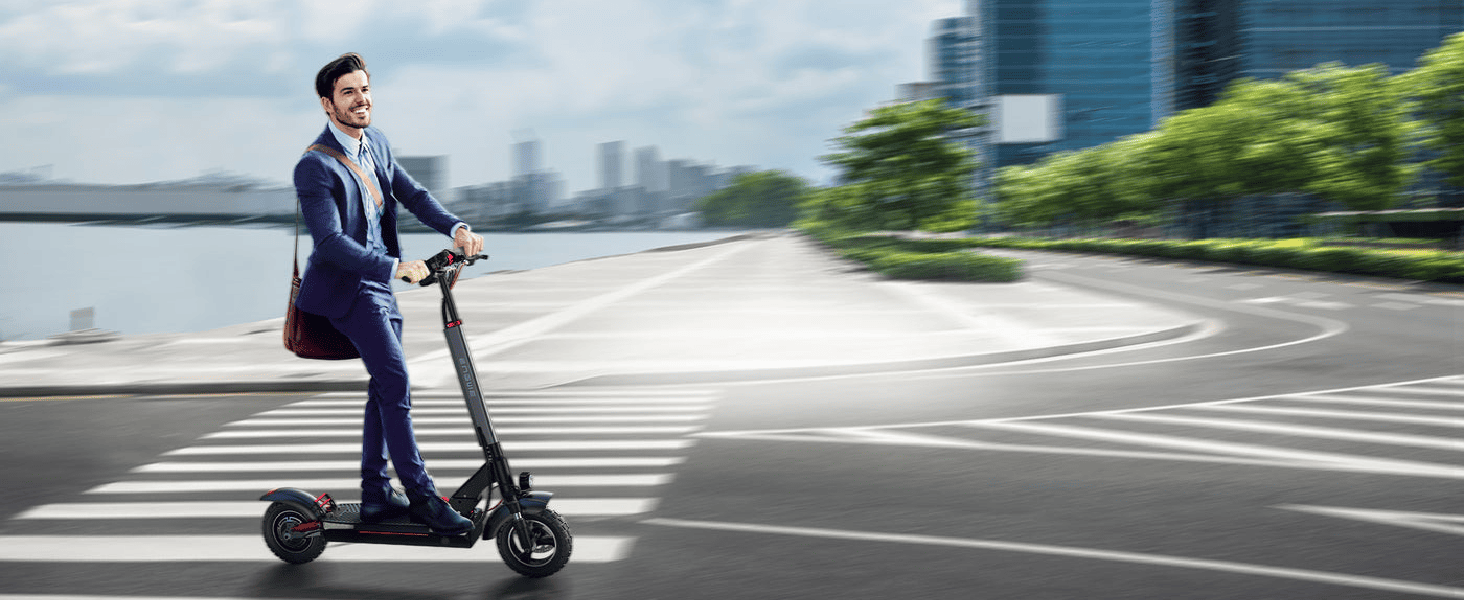 Businessman in a suit riding an electric scooter on a city road with trees and buildings in the background.