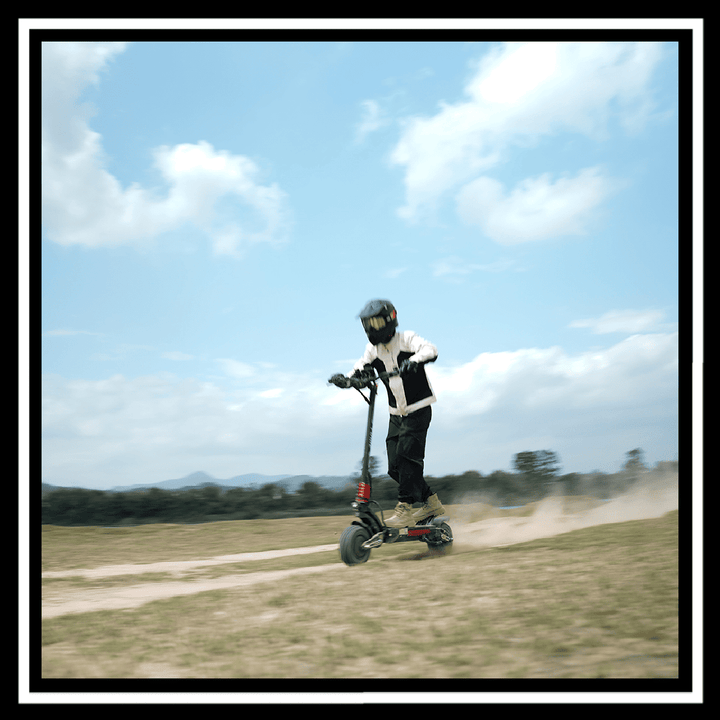 Person riding the YUME Raptor 2 electric scooter on a dirt path in a field under a clear blue sky.