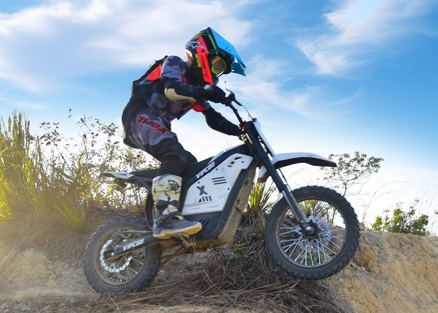 A rider in protective gear jumps a dirt hill on an electric dirt bike against a blue sky.