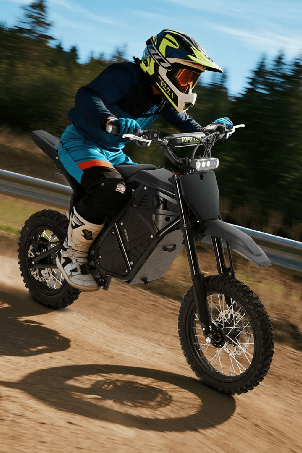 A rider in a blue outfit and helmet speeds on an electric dirt bike on a dirt trail surrounded by trees.