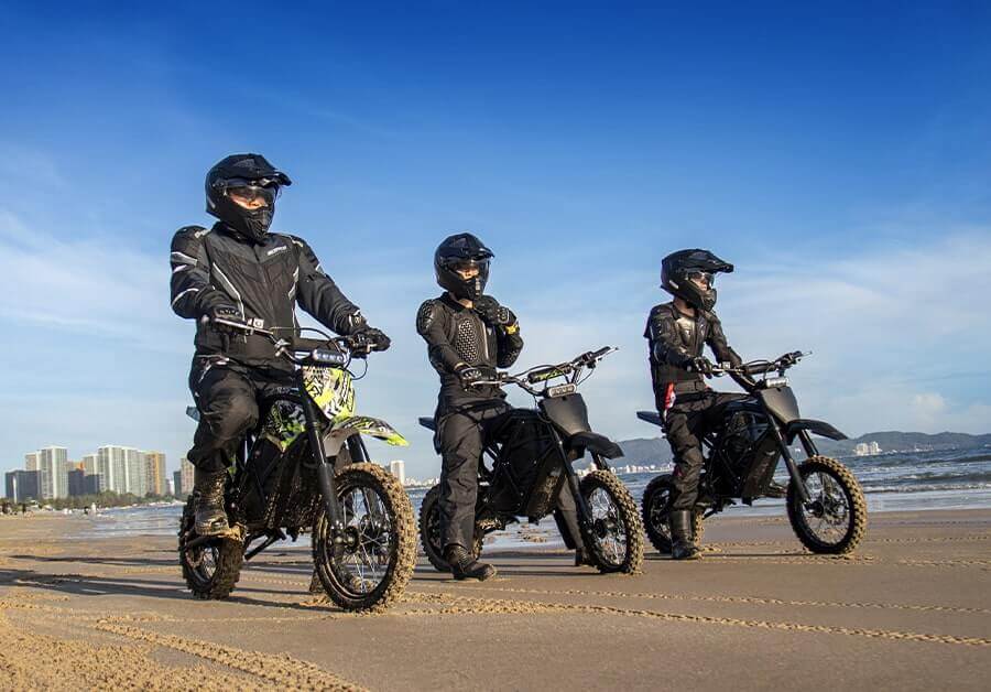 Three riders on dirt bikes enjoying a beach ride with a city skyline in the background.