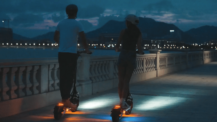 Couple riding the 🇮🇪🇪🇺 KUKIRIN M4 MAX scooters at night along a scenic waterfront path.
