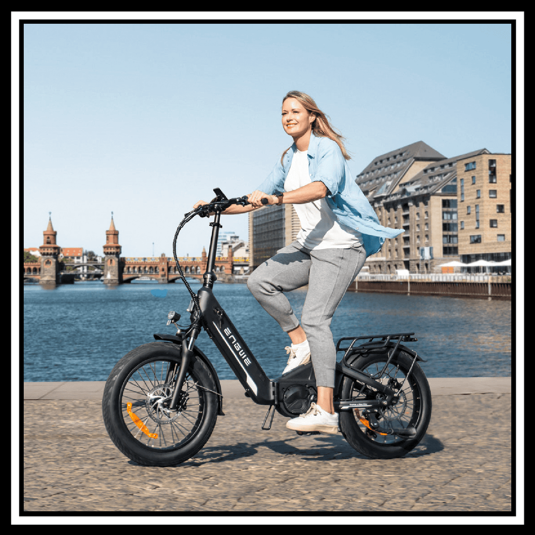 Woman riding the 🇮🇪🇬🇧🇪🇺 ENGWE L20 3.0 PRO electric bike by the water with a city backdrop.