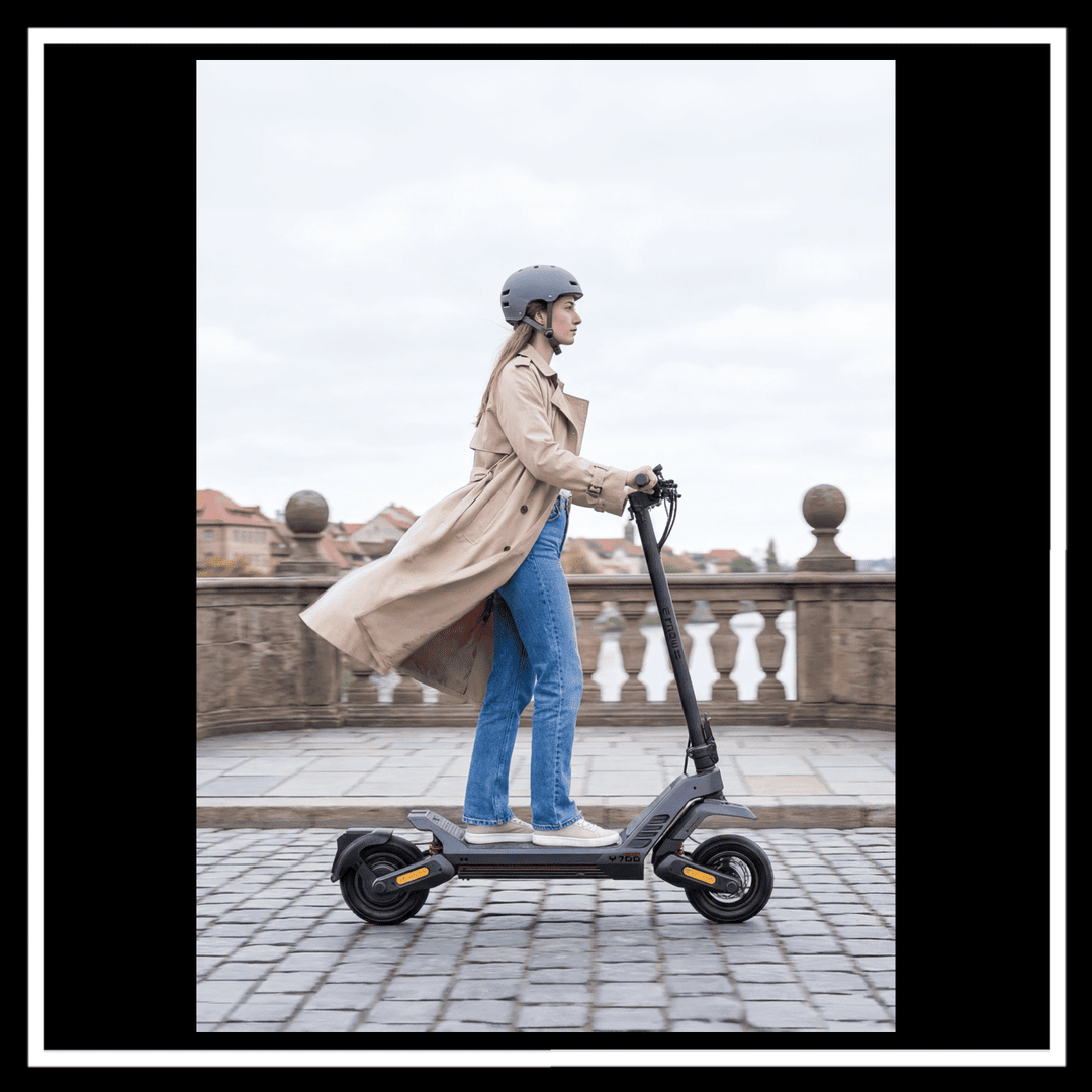 Woman riding ENGWE Y700 Electric Scooter on cobblestone path, wearing a helmet and trench coat.