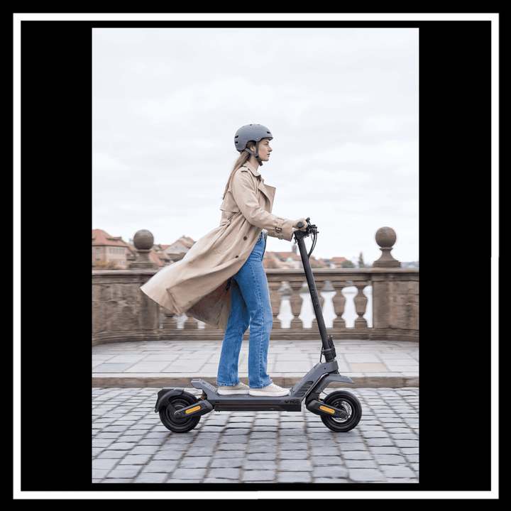 Woman riding ENGWE Y700 Electric Scooter on cobblestone path, wearing a helmet and trench coat.