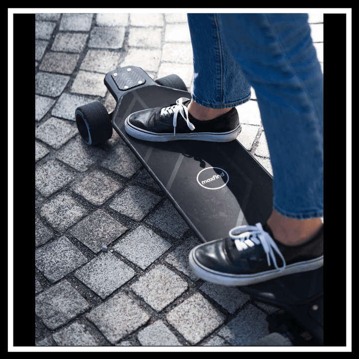 Person riding a Maxfind MAX6 electric skateboard on a cobblestone path.