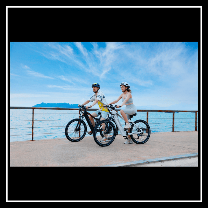 Two cyclists riding SAMEBIKE XD26-II electric bikes along a scenic waterfront promenade.