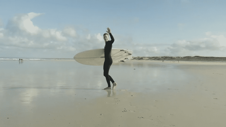 Surfer in wetsuit walking on the beach, holding a surfboard and waving at the camera.