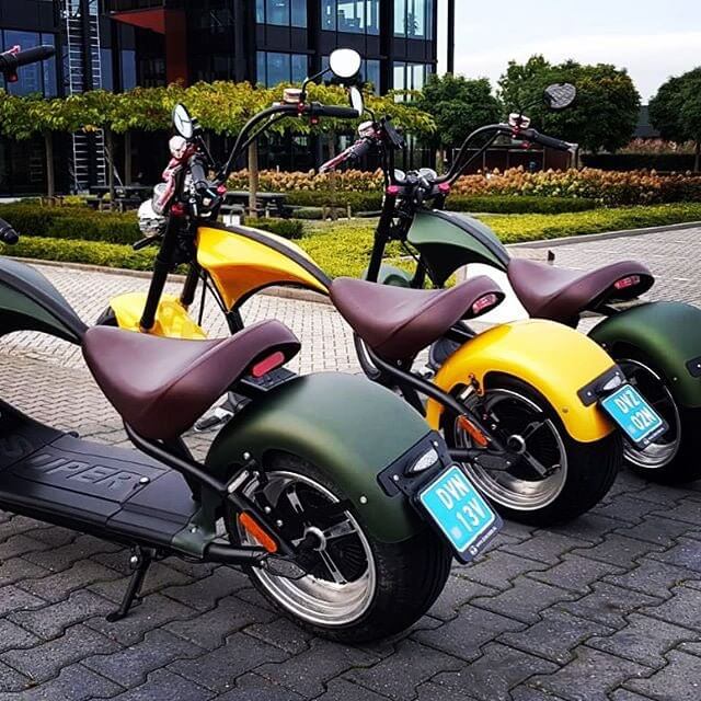 M1P electric chopper motorcycle in green, yellow, and black parked on a paved area with greenery in the background.