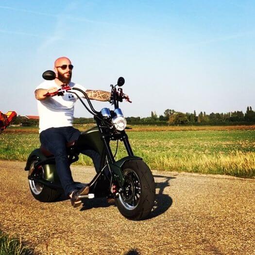 M1P electric chopper motorcycle being ridden on a rural road with fields in the background.