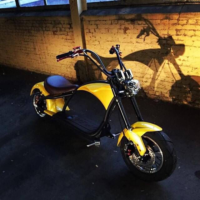 M1P electric chopper motorcycle in yellow color positioned against a brick wall, highlighting its sleek design.