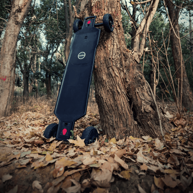 Maxfind FF AT electric skateboard leaning against a tree in a forest with fallen leaves.